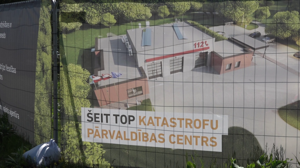 Būvlaukuma plakāts ar uzrakstu "ŠEIT TOP KATASTROFU PĀRVALDĪBAS CENTRS" un ēkas vizualizāciju