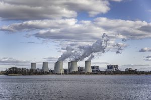 Ainava ar dūmojošiem industriāliem skursteņiem pie ūdens un mākoņainām debesīm