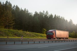 Sarkans kravas auto brauc pa šoseju gar mežu