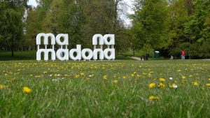 Madonas parka pļava ar uzrakstu “madona” un pienenēm priekšplānā