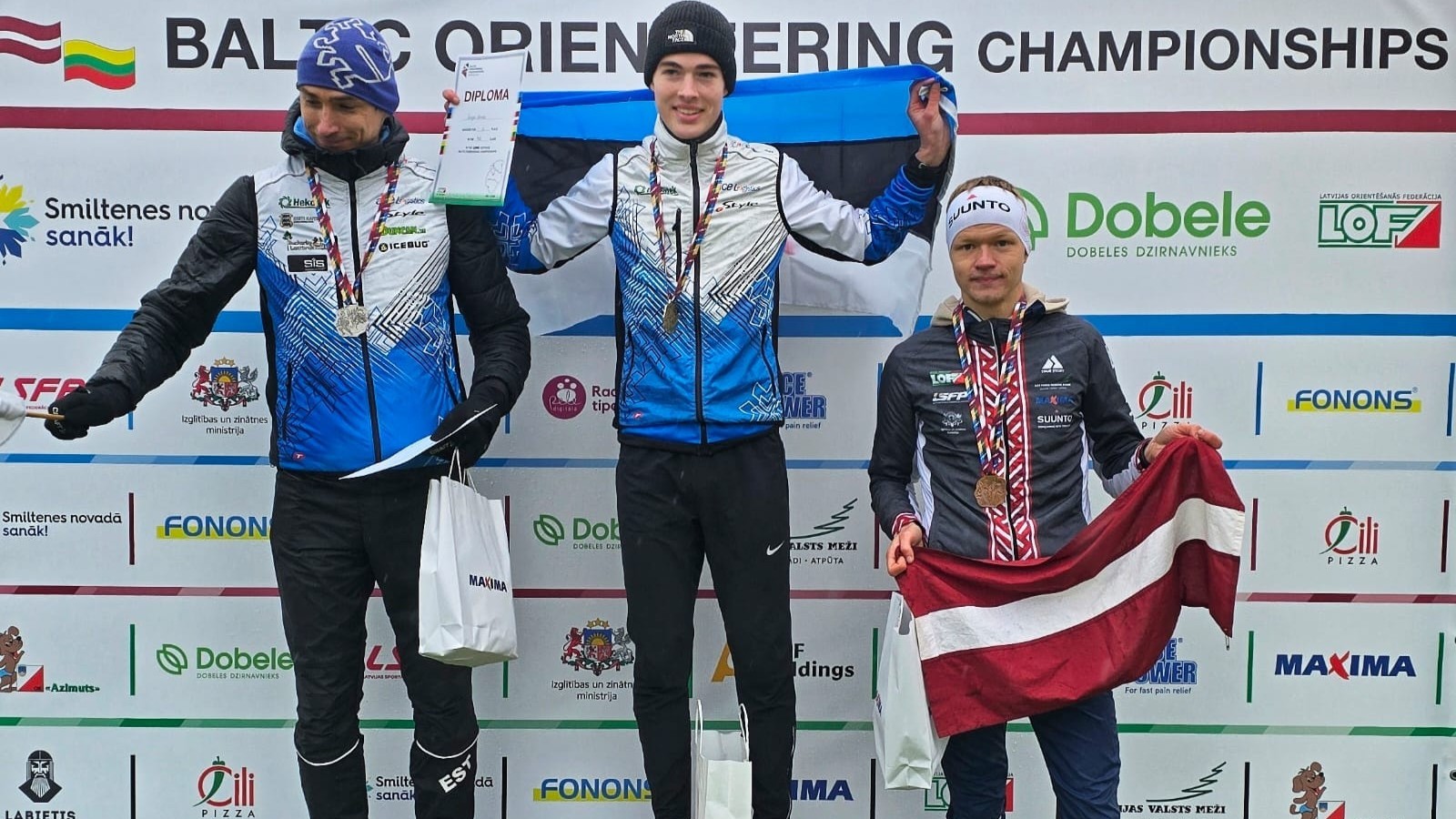 Trīs orientēšanās sportisti ar medaļām un diplomiem stāv uz pjedestāla Baltijas čempionātā vīriešiem