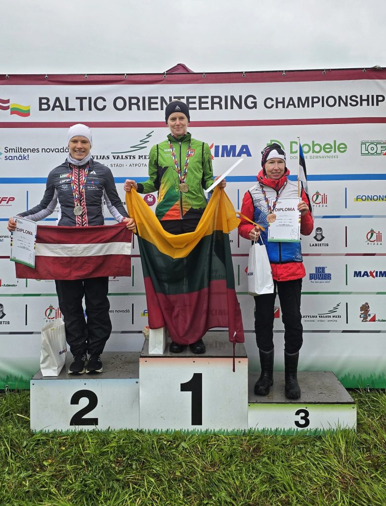 Trīs orientēšanās sportistes ar medaļām un diplomiem stāv uz pjedestāla Baltijas čempionātā.