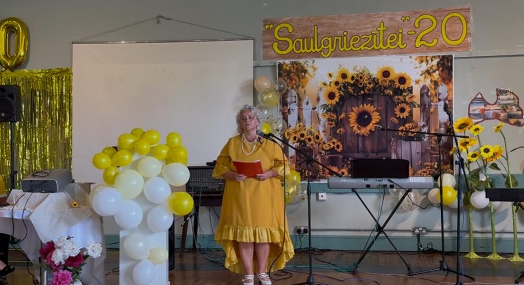 Sieviete dzeltenā kleitā stāv uz skatuves pie mikrofona, runājot vai uzstājoties "Saulgriezīte" 20 gadu jubilejas pasākumā
