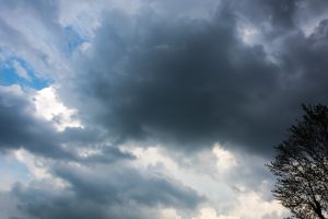 Tumši lietus mākoņi debesīs ar daļēji redzamu koku siluetu