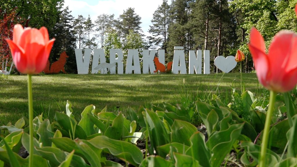 Uzraksts “Varakļāni” parkā ar sarkanām tulpēm priekšplānā un mežu fonā