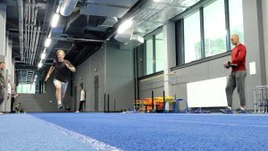Sportists veic sprinta testu iekštelpu vieglatlētikas hallē, trenerim vērojot rezultātus.