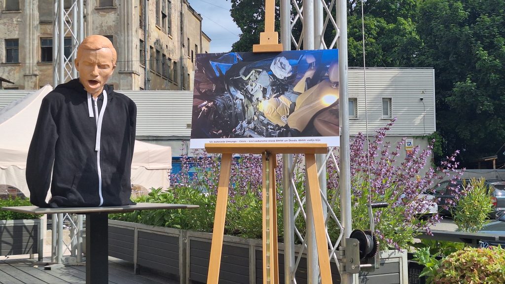 Manekens melnā sporta jakā stāv blakus molbertam ar lielu fotogrāfiju, kas, iespējams, attēlo satiksmes negadījuma sekas