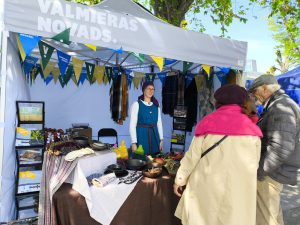 Tirdziņa stends ar uzrakstu "Valmieras novads" Hanzas dienās, kur pārdevēja tradicionālā tērpā sarunājas ar pircējiem, piedāvājot amatniecības izstrādājumus