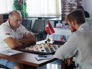 Divi dambretisti spēlē dambreti turnīra laikā, redzams Latvijas karogs