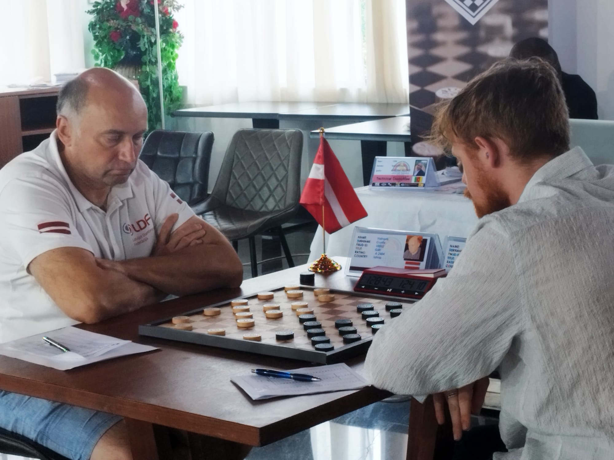 Divi dambretisti spēlē dambreti turnīra laikā, redzams Latvijas karogs