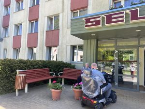 Seniori pie pansionāta “Selga” ieejas sēž uz soliņa un pārvietojas ar elektrisko skūteri