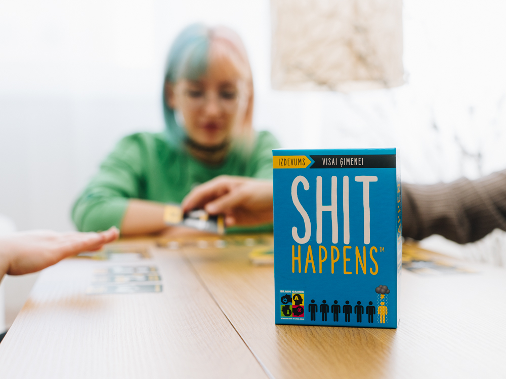 Spēles "Shit Happens" kaste uz galda ar cilvēkiem fonā