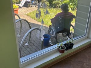 Seniors ratiņkrēslā sēž pansionāta dārzā, skats caur logu