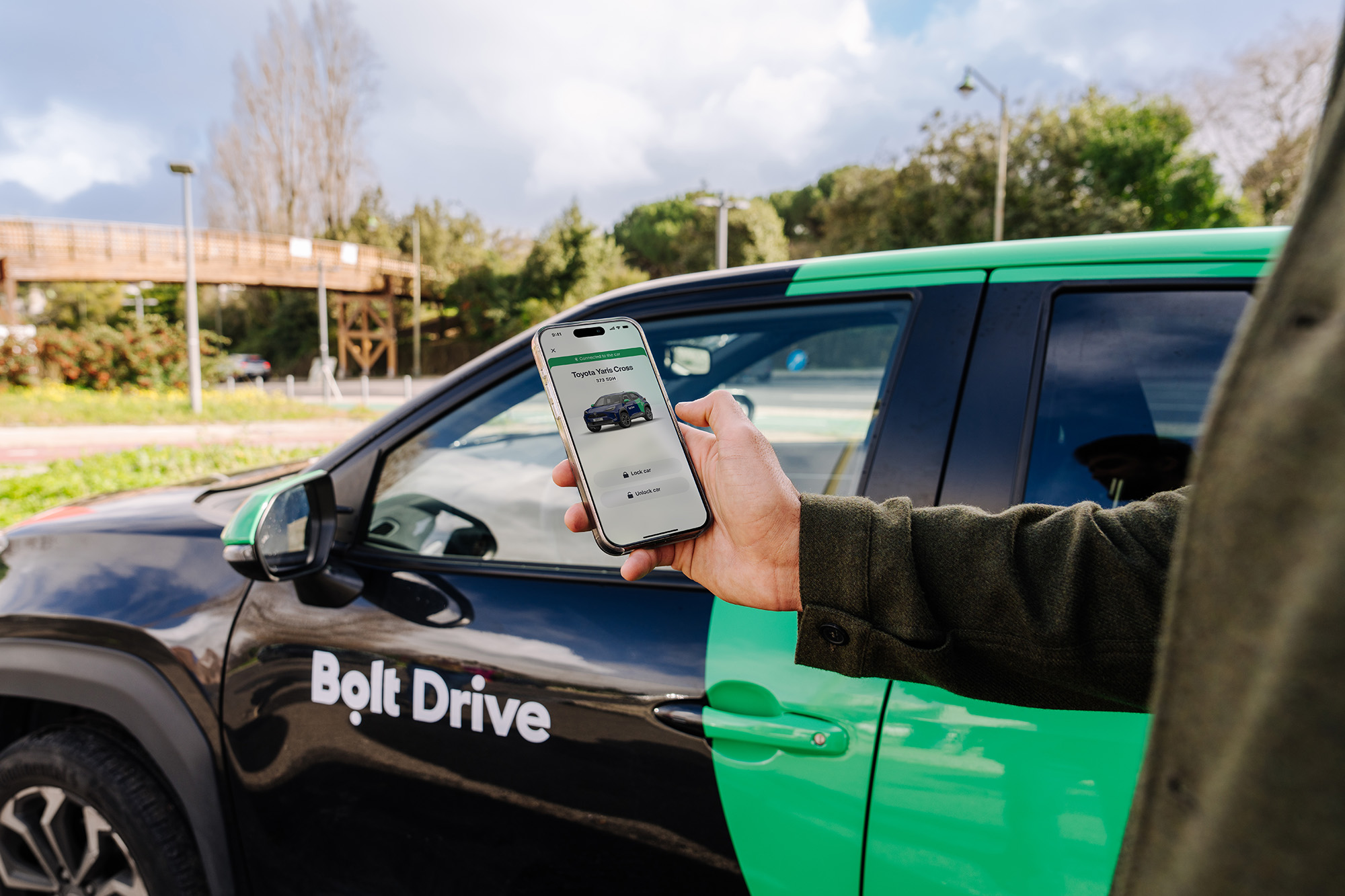 Persona, kas ar viedtālruni atver Bolt Drive automašīnu, kurai ir melns un zaļš dizains ar 