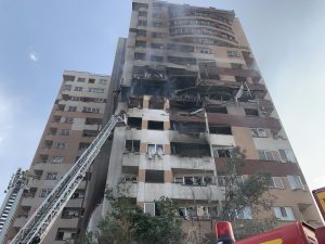 Daudzdzīvokļu ēka pēc sprādziena ar redzamiem bojājumiem un ugunsdzēsējiem uz izlices