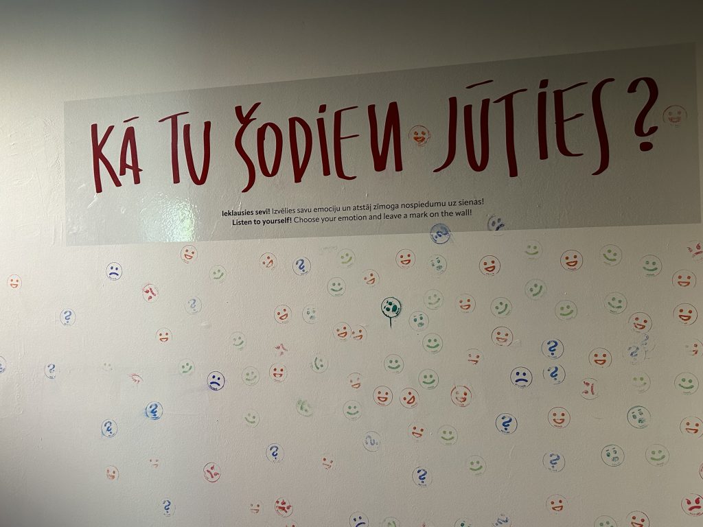 Siena ar uzrakstu "KĀ TU ŠODIEN JŪTIES?" un daudziem zīmogotiem smaidiņiem un jautājuma zīmēm