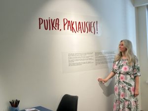 Sieviete ar blondiem matiem un kleitu stāv pie baltas sienas ar uzrakstu "PUIKA, PAKLAUSIES!" un skaidrojošu tekstu