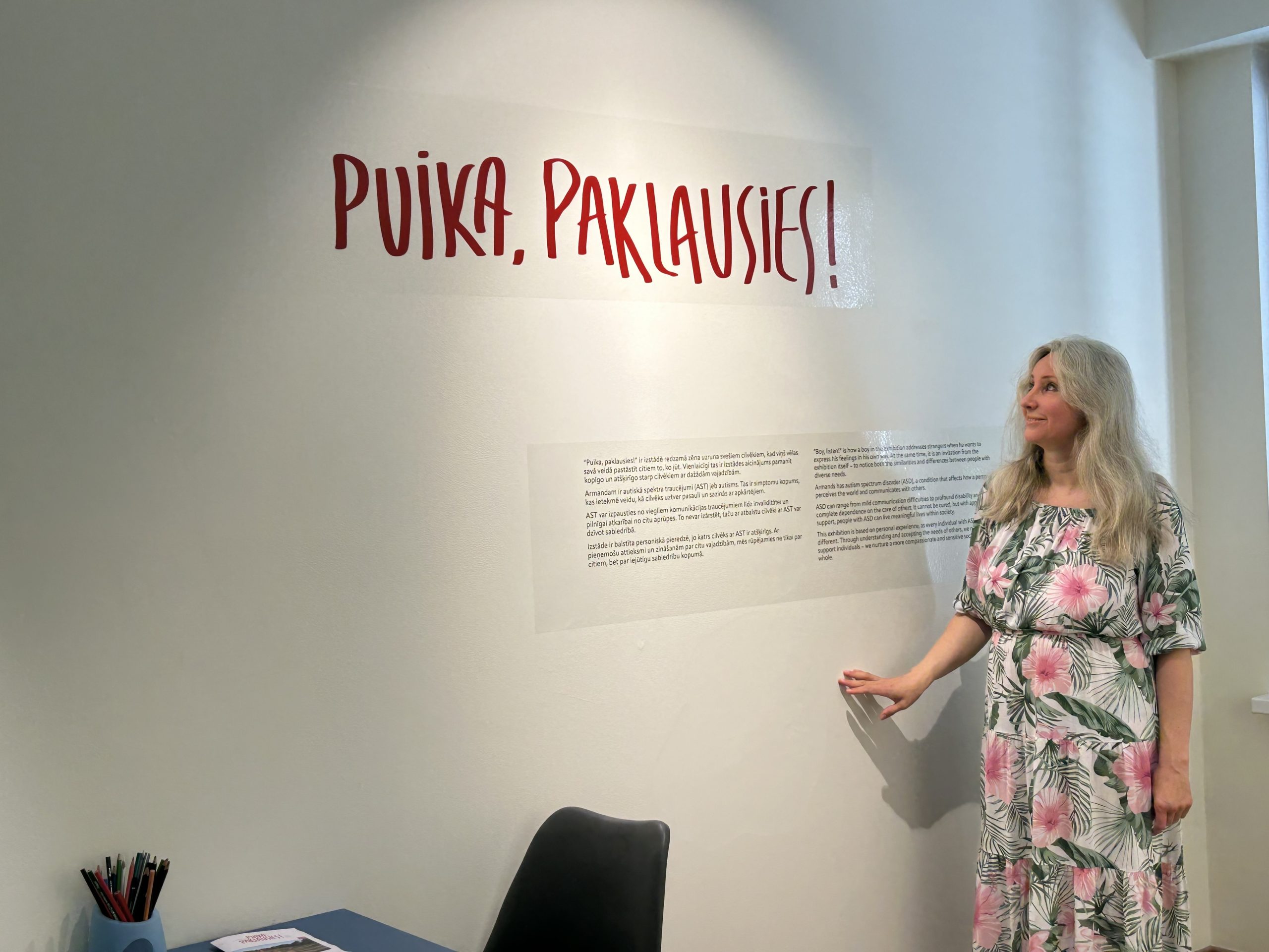 Sieviete ar blondiem matiem un kleitu stāv pie baltas sienas ar uzrakstu 