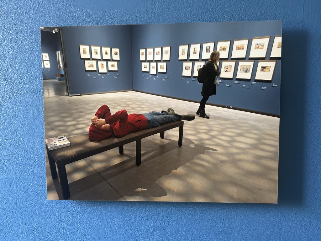 Fotoattēls izstādē, kurā zēns sarkanā jakā guļ uz soliņa, kamēr cita persona apskata gleznas galerijā
