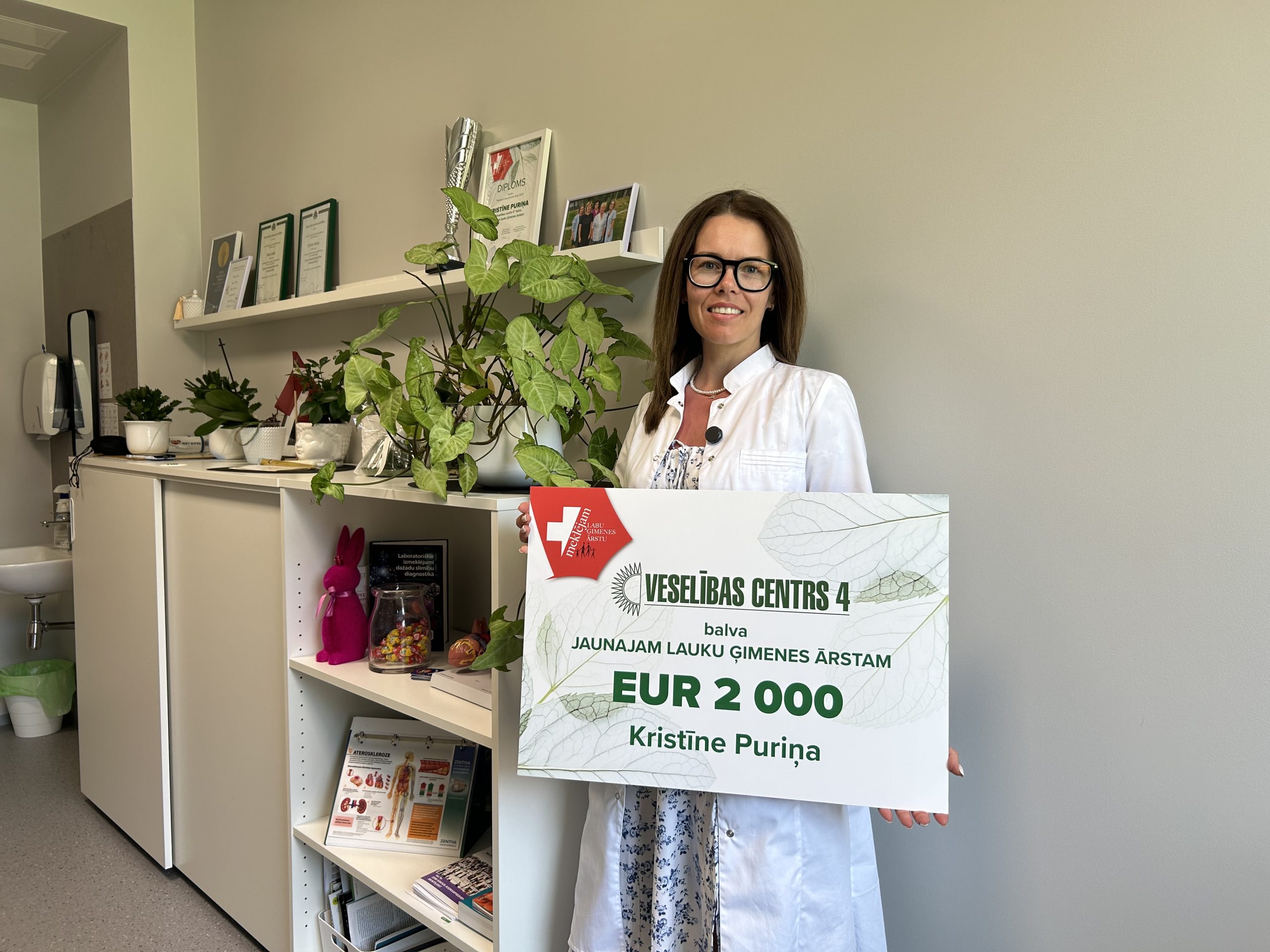 Jauna ģimenes ārste, Kristīne Puriņa, baltā halātā stāv birojā un tur rokās lielu simbolisku čeku ar uzrakstu 