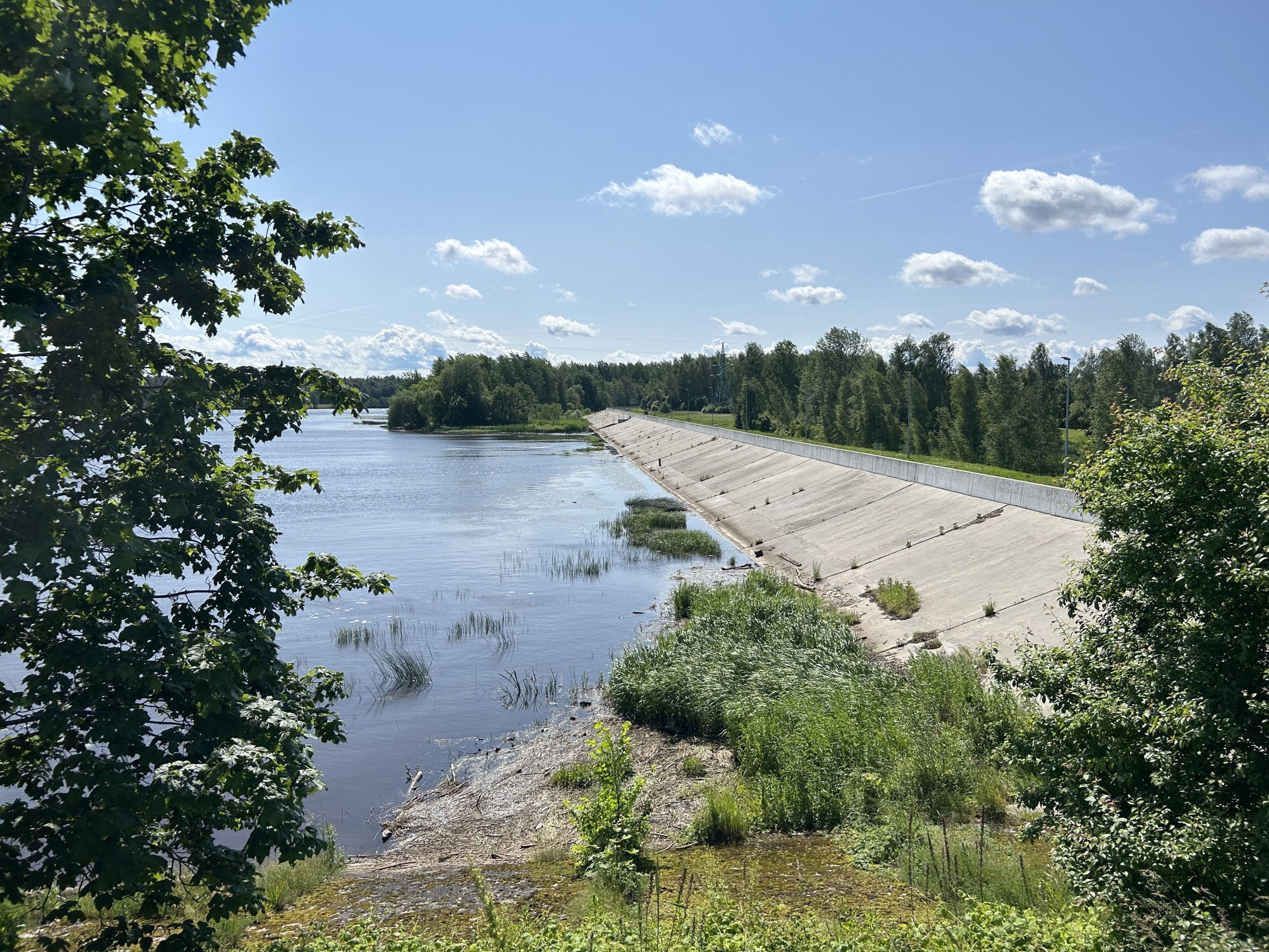 Panorāmas skats uz Daugavu ar zaļu veģetāciju un betona konstrukciju gar krastu