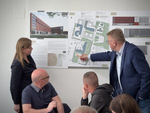 Cilvēki apspriež Rēzeknes slimnīcas fasādes atjaunošanas projektu, rādot uz ēkas plānu