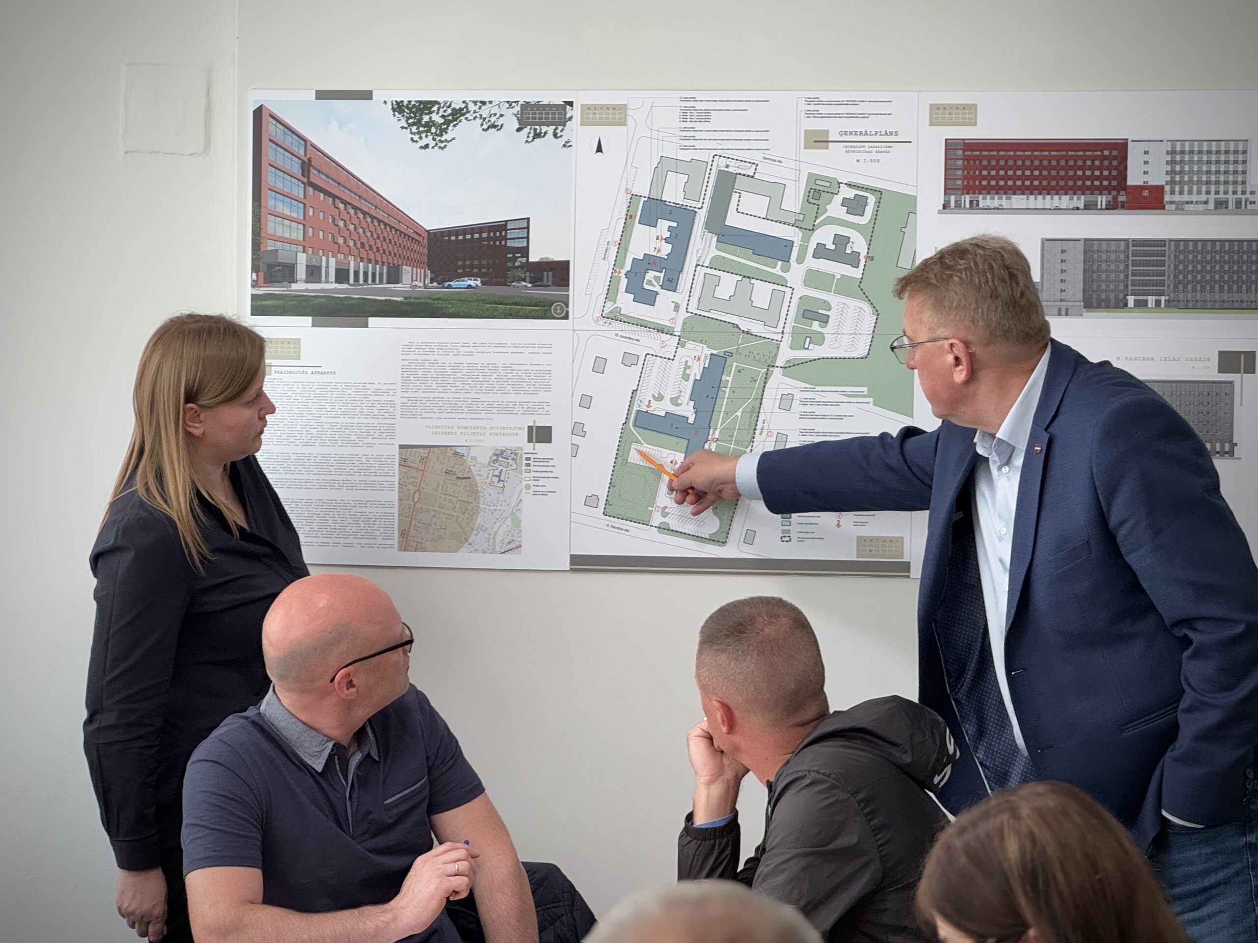 Cilvēki apspriež Rēzeknes slimnīcas fasādes atjaunošanas projektu, rādot uz ēkas plānu