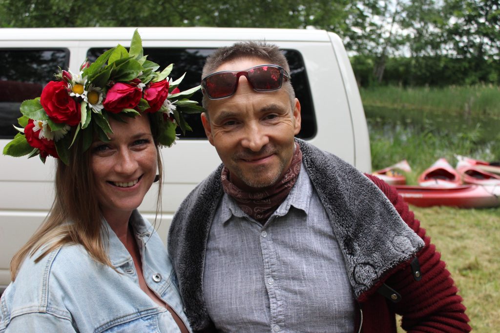 Ingūna Pranka ar Jāņu vainagu galvā un Atis Luguzs saulesbrillēs, stāvot blakus un smaidot kamerā, ar baltu busiņu un sarkanām laivām fonā