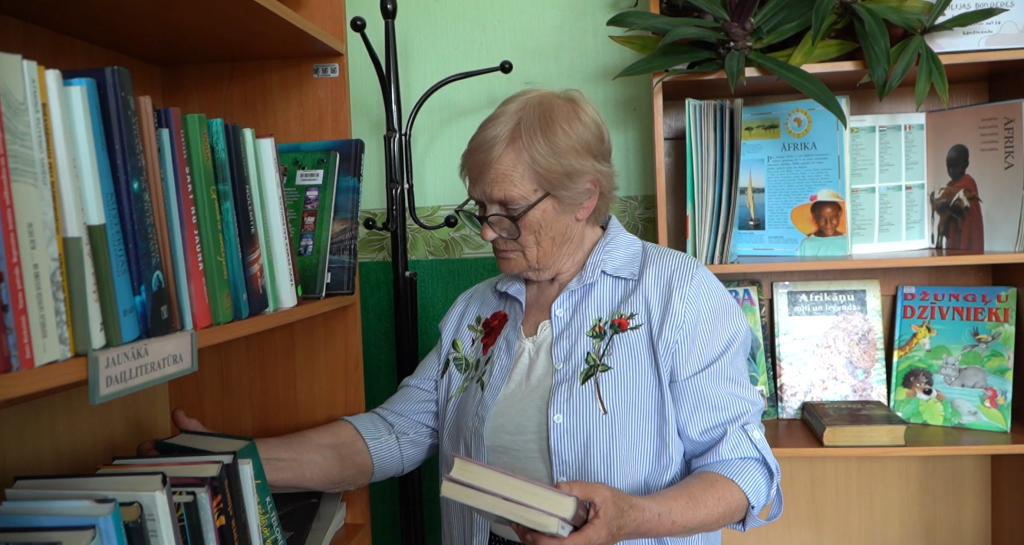 anīna Blūzma bibliotēkā pēta grāmatu plauktu un tur rokās grāmatu