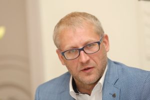 Vīrietis ar gaišiem matiem un brillēm, uzvalkā, skatās tieši kamerā ar nopietnu sejas izteiksmi