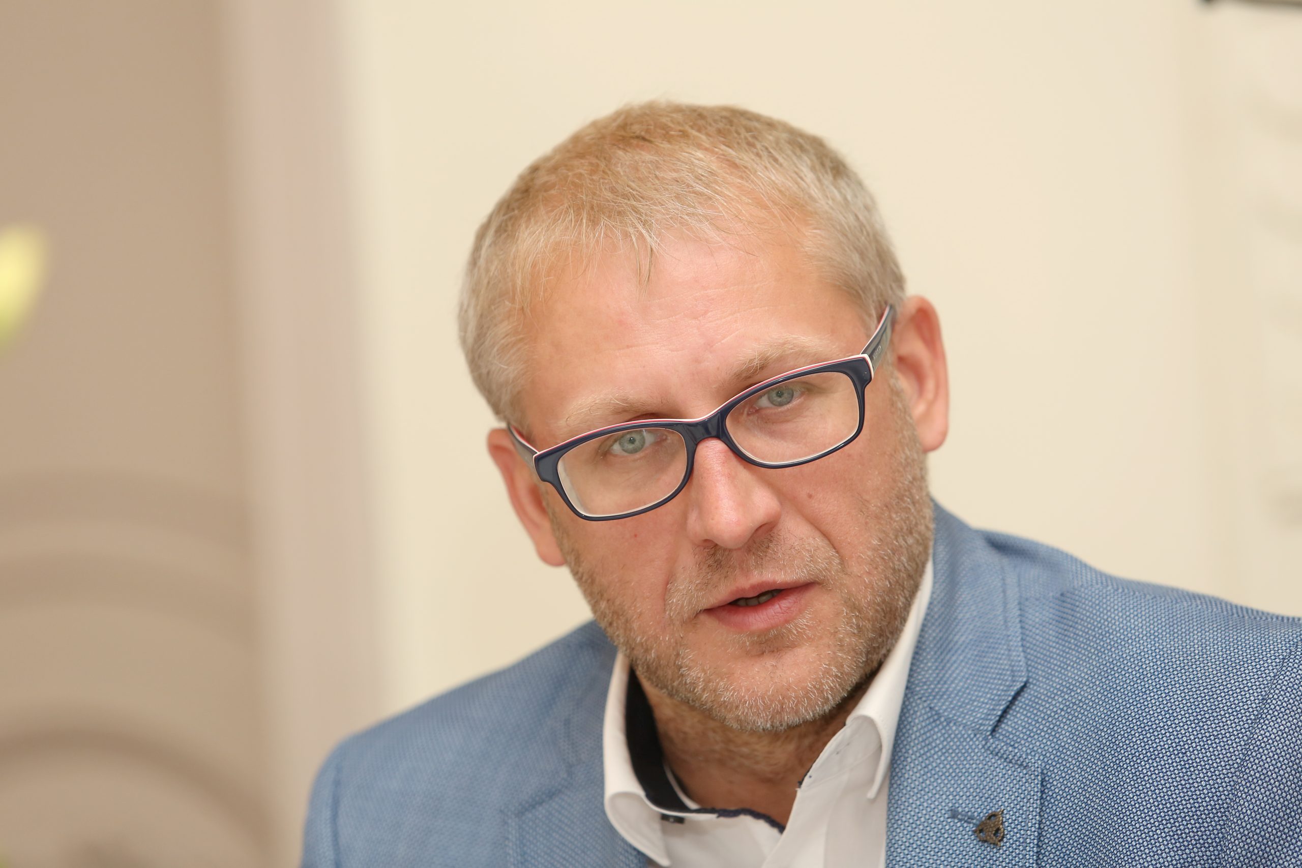 Vīrietis ar gaišiem matiem un brillēm, uzvalkā, skatās tieši kamerā ar nopietnu sejas izteiksmi