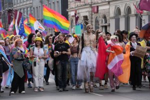 Cilvēku grupa ar varavīksnes karogiem un dažādiem tērpiem dodas gājienā pa ielu "Riga Pride 2024" laikā