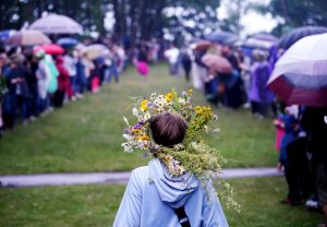 Cilvēks ar Jāņu vainagu uz galvas lietainās Jāņu svinībās, kas stāv zālājā starp cilvēku pūli ar lietussargiem