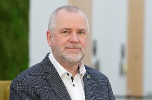 Vīrietis ar sirmu bārdu un uzvalkā, smaida un skatās tieši kamerā.