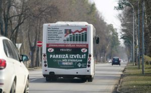 Autobusa aizmugure ar politisko reklāmu uz ceļa, kas ved prom no skatītāja, ar kokiem abās pusēs