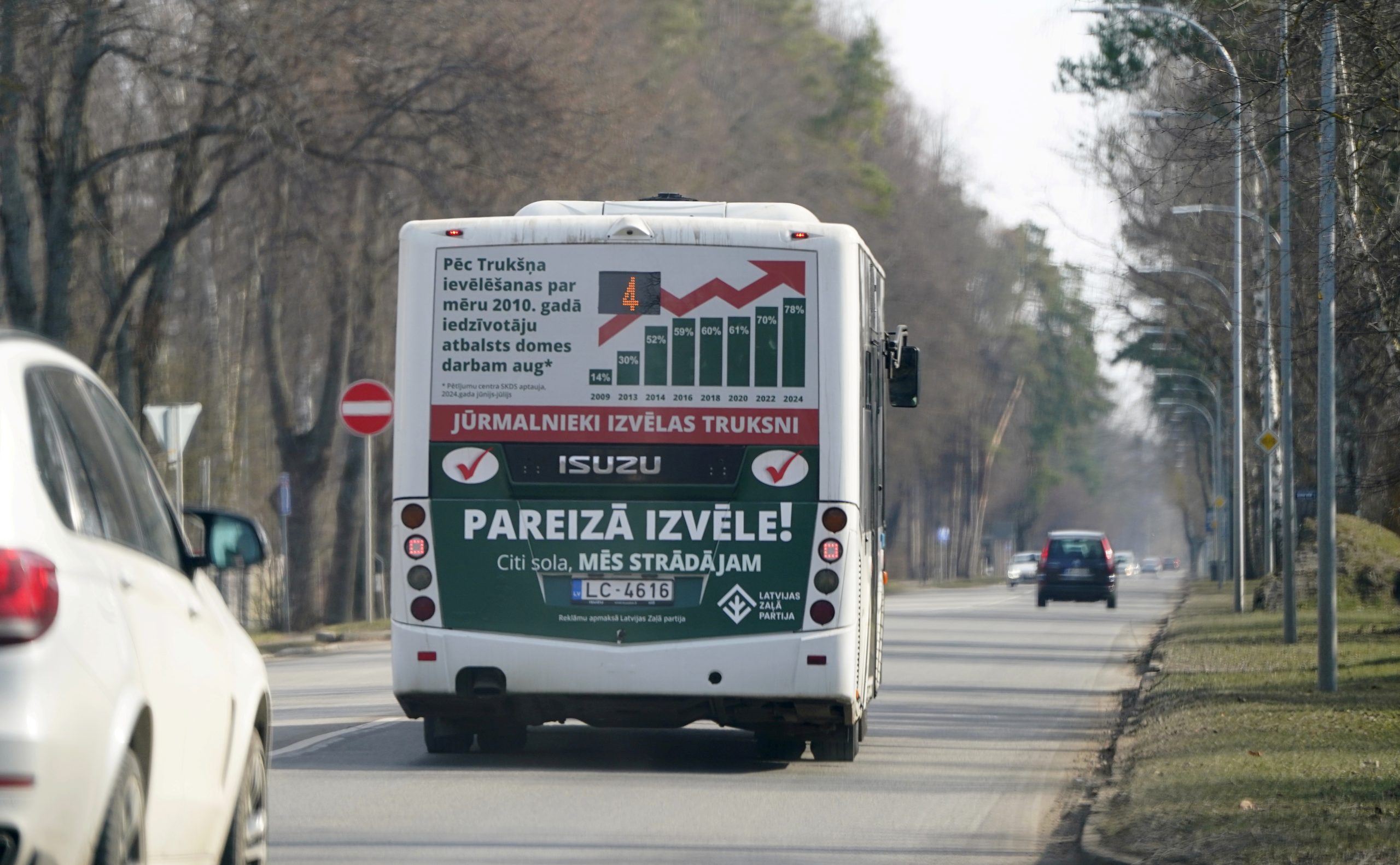 Autobusa aizmugure ar politisko reklāmu uz ceļa, kas ved prom no skatītāja, ar kokiem abās pusēs
