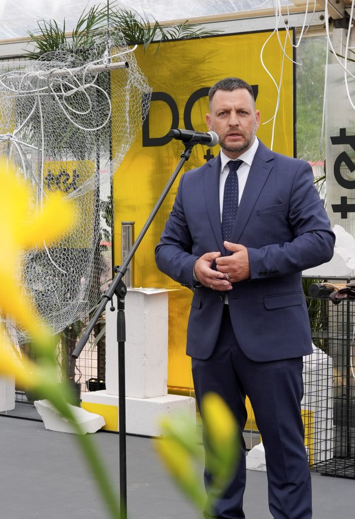Vīrietis tumšā uzvalkā runā pie mikrofona uz skatuves ar dzeltenu fonu un dekorācijām