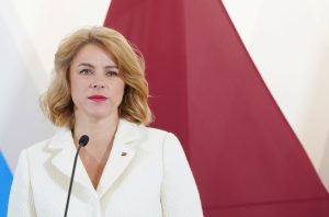 Ministru prezidente Evika Siliņa baltā žaketē pie mikrofona ar Latvijas karoga fonu