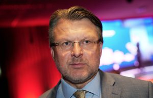 Ainārs Šlesers stāv konferencē uz sarkani zilas gaismas fona.