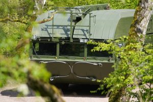 Militārais transportlīdzeklis mežā aizsegts ar koku zariem