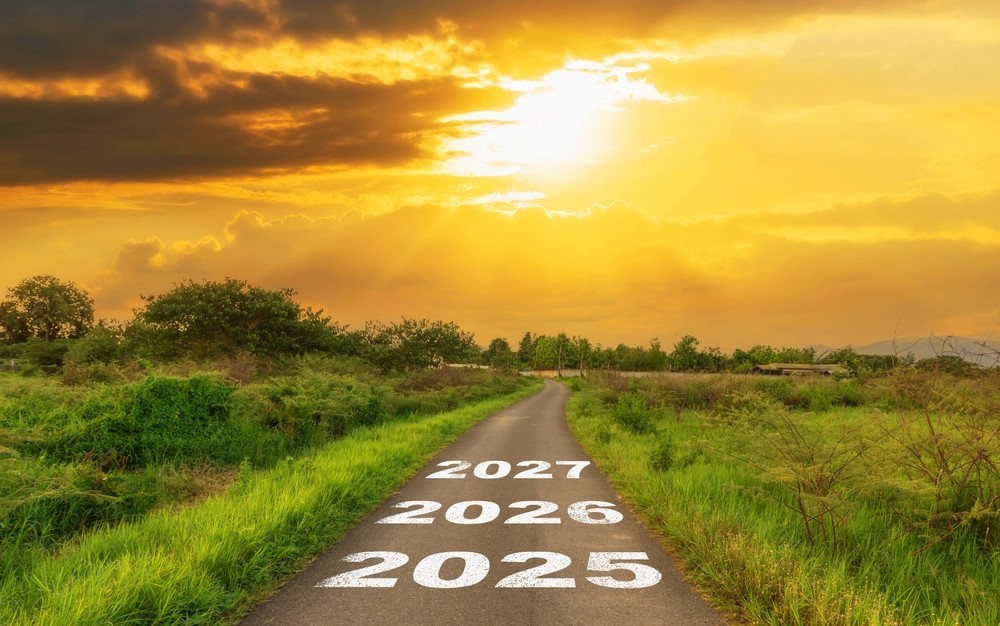Ceļš lauku ainavā ar uzrakstiem “2025”, “2026”, “2027” ved uz horizontu saulrietā – simbolizē plānu nākotnei reto slimību jomā.
