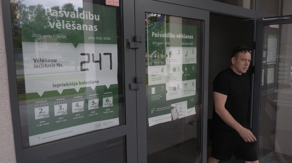 Vīrietis melnā apģērbā iziet no ēkas, kuras logā ir izvietots vēlēšanu plakāts ar informāciju par Pašvaldību vēlēšanām 2025. gada 7. jūnijā.