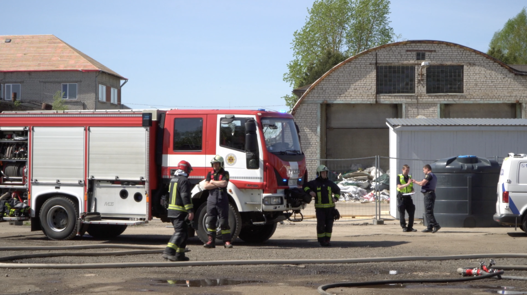 Ugunsdzēsēji un citi dienestu darbinieki stāv pie liela sarkana ugunsdzēsēju auto ar baltām un sarkanām svītrām uz sāna, pie vecām ēkām