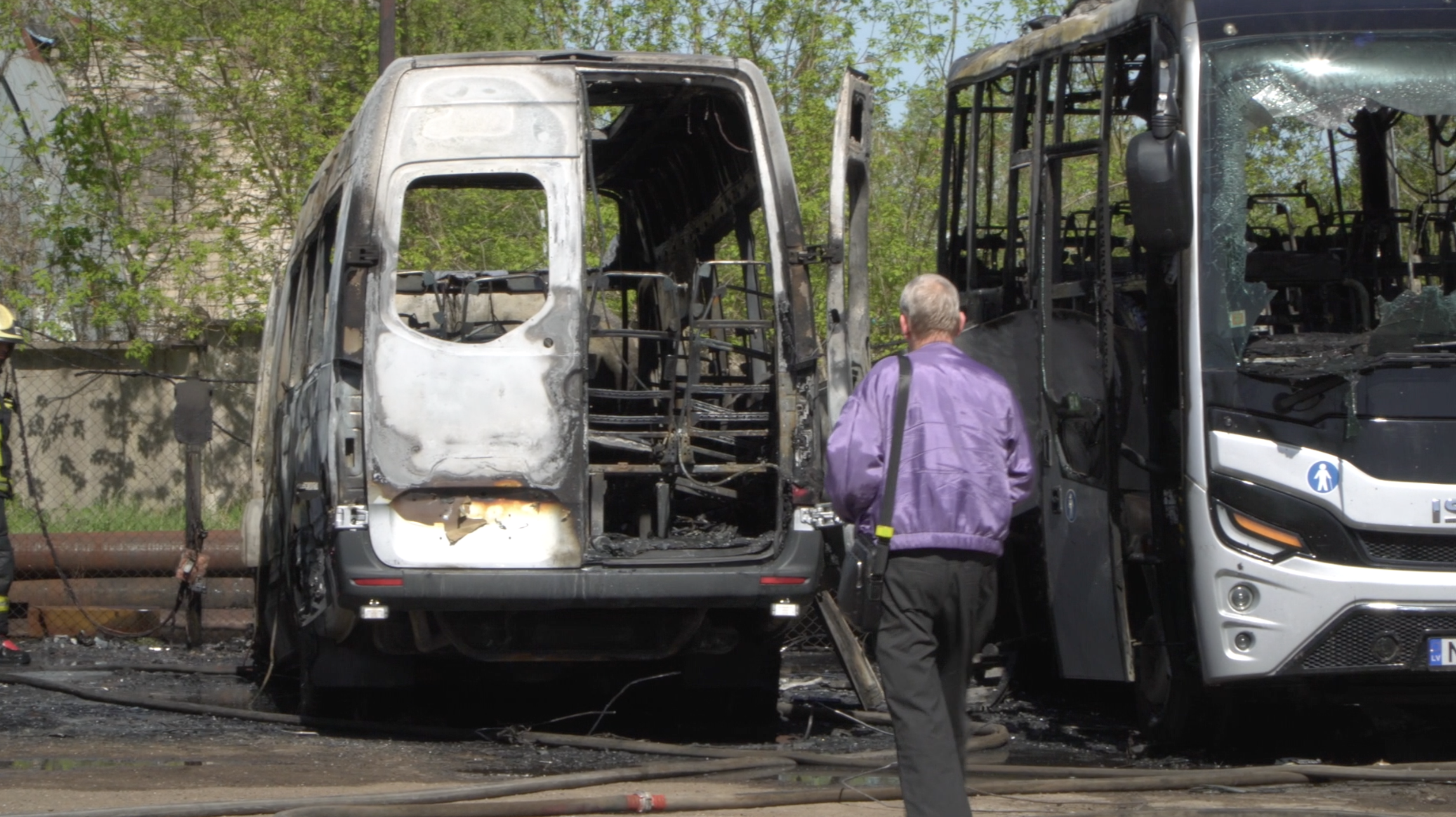 Izdedzis balts mikroautobuss un melns autobuss stāv blakus, vīrietis violetā jakā skatās uz bojātajiem transportlīdzekļiem