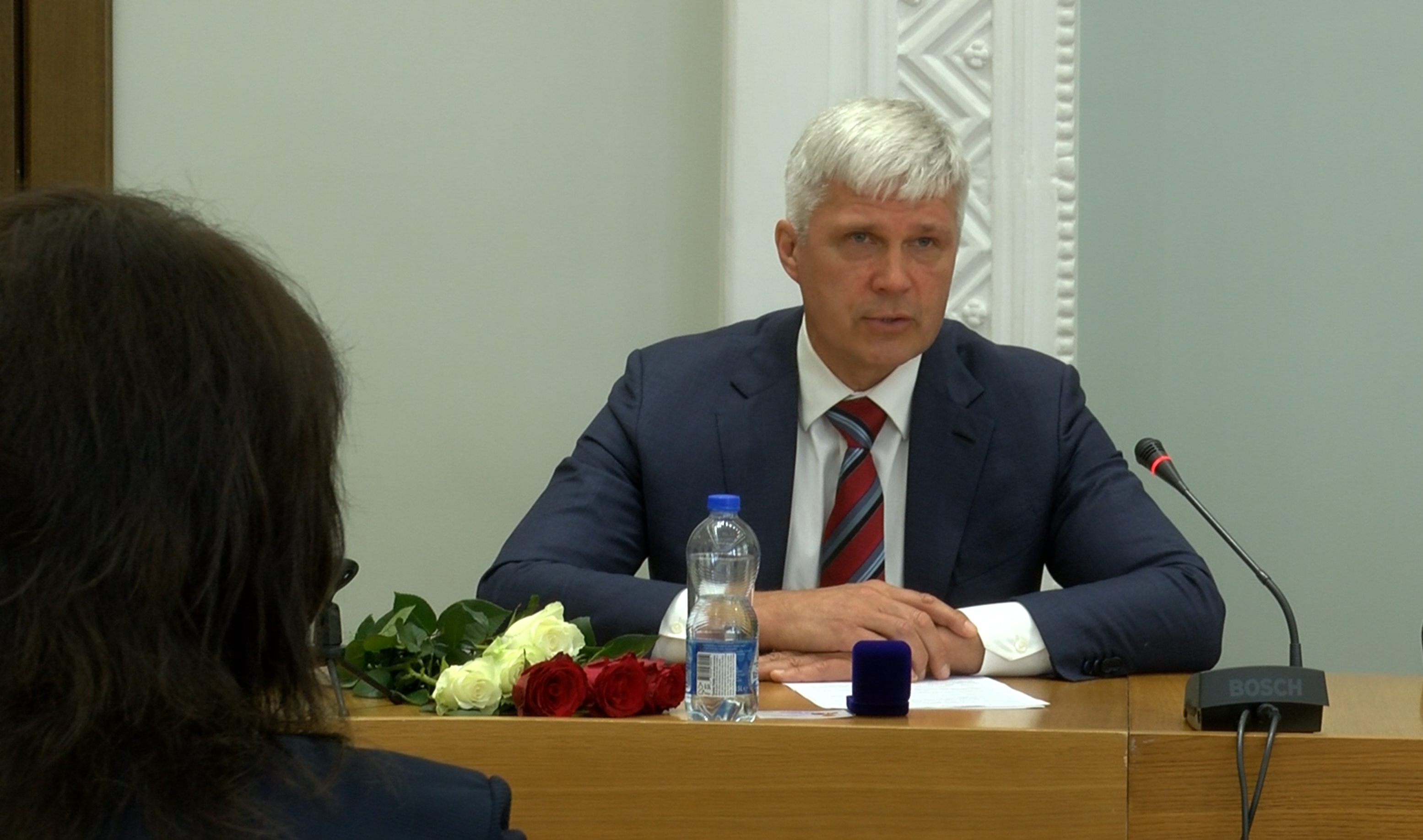 Aleksandrs Bartaševics uzrunā domes sēdē, pie galda ar rozēm un mikrofonu