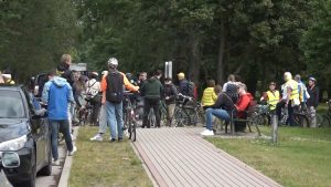 Grupa cilvēku ar velosipēdiem pulcējas pie ceļa un zaļas zonas