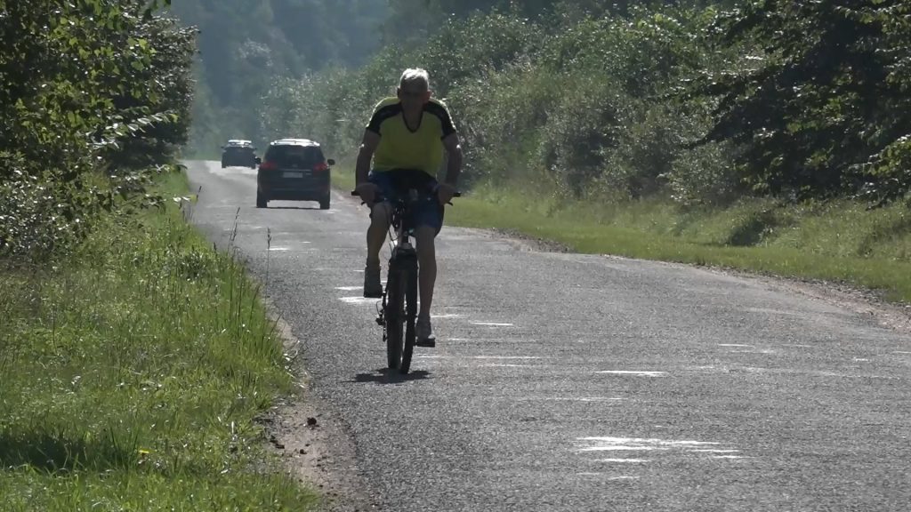 Vīrietis brauc ar velosipēdu pa asfaltētu ceļu, fonā redzamas automašīnas