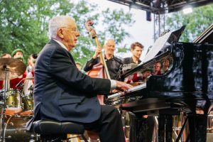 Maestro Raimonds Pauls uzstājas uz skatuves Tigulkalna koncertā, fonā redzami mūziķi un koristi tautastērpos