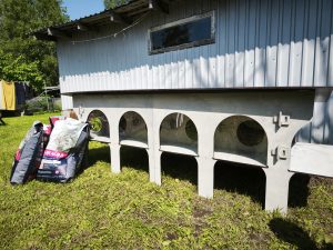 Kaķu mājiņa ar vairākiem nodalījumiem un kaķu barības maisiem pagalmā saulainā dienā