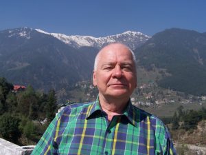 Jānis Zaļkalns stāv kalnu ainavas fonā ar sniegotām virsotnēm un zaļām nogāzēm